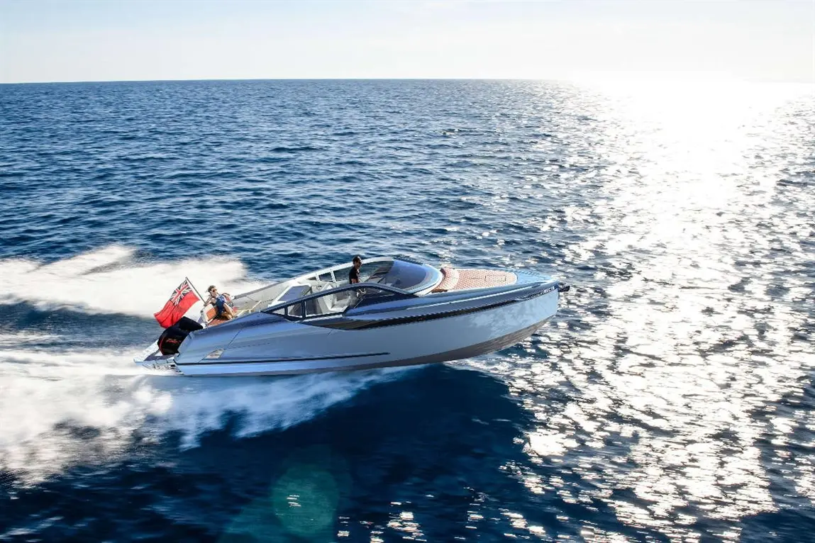 A sleek motorboat speeds across the ocean, with a person waving a British flag and another inside the boat.